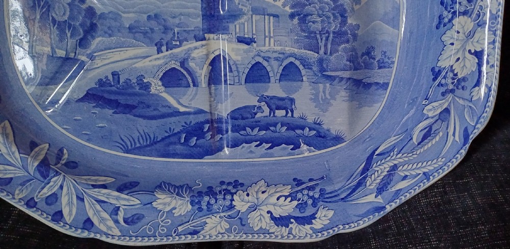 An oval blue and white transfer-printed earthenware meat platter depicting a central landscape with a bridge, tower, and cattle framed by a broad vine leaf border. View VII