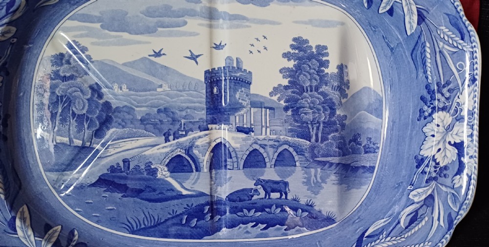 An oval blue and white transfer-printed earthenware meat platter depicting a central landscape with a bridge, tower, and cattle framed by a broad vine leaf border. View VIII