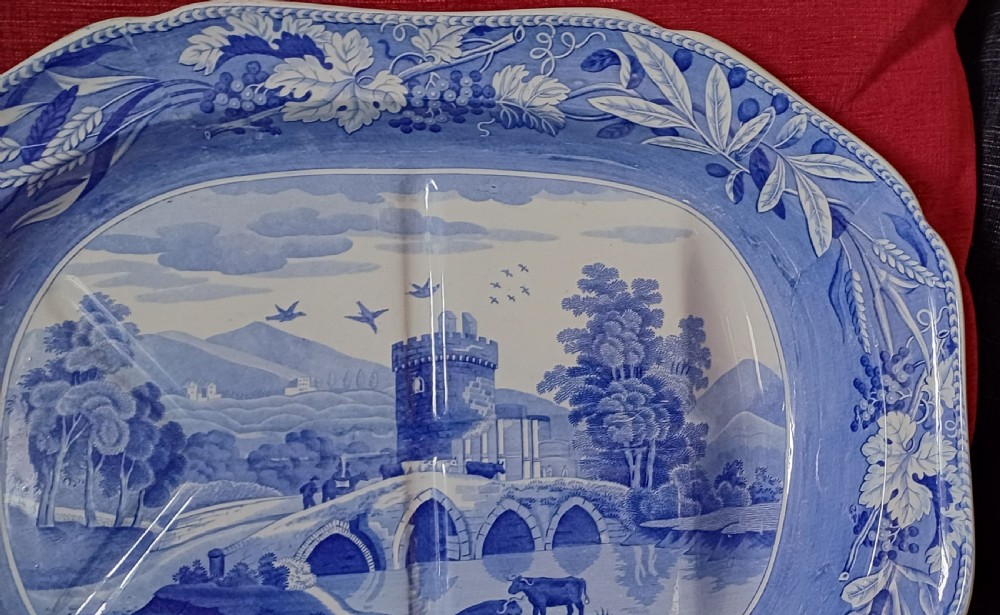 An oval blue and white transfer-printed earthenware meat platter depicting a central landscape with a bridge, tower, and cattle framed by a broad vine leaf border. View IX