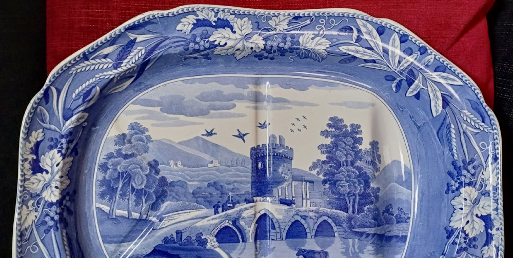 An oval blue and white transfer-printed earthenware meat platter depicting a central landscape with a bridge, tower, and cattle framed by a broad vine leaf border. View X