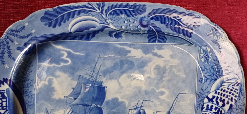 An oval blue and white earthenware transferware platter featuring a central naval sea battle scene surrounded by a botanical and seashell border. View III