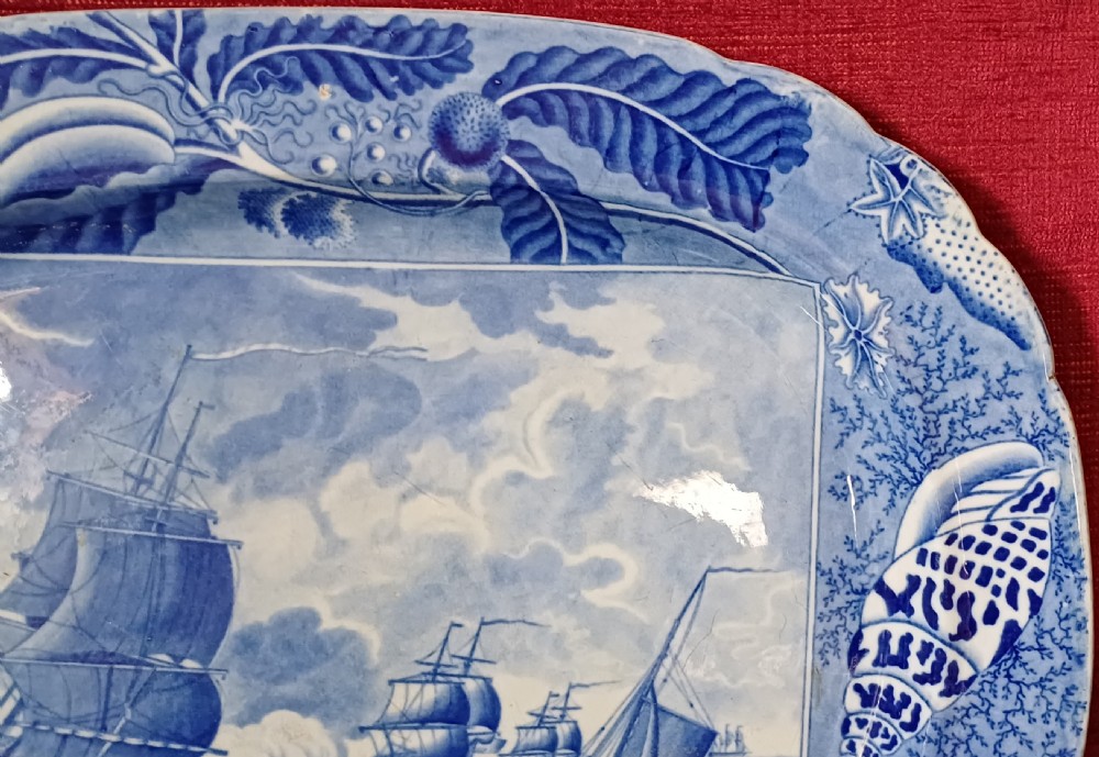 An oval blue and white earthenware transferware platter featuring a central naval sea battle scene surrounded by a botanical and seashell border. View IV