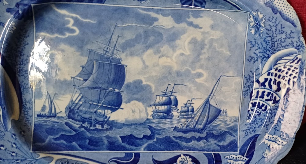 An oval blue and white earthenware transferware platter featuring a central naval sea battle scene surrounded by a botanical and seashell border. View VI