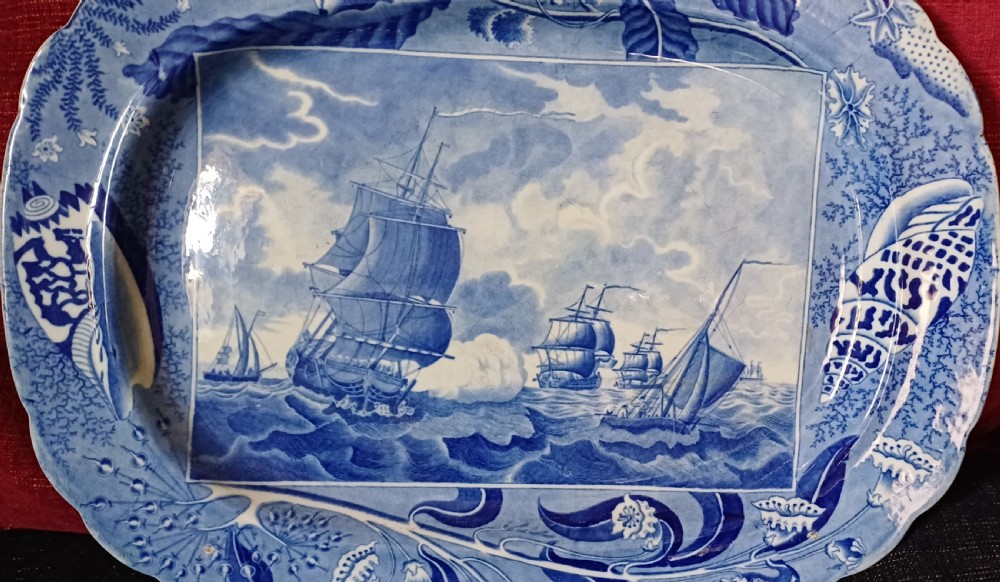An oval blue and white earthenware transferware platter featuring a central naval sea battle scene surrounded by a botanical and seashell border. View VII