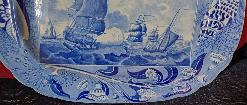 An oval blue and white earthenware transferware platter featuring a central naval sea battle scene surrounded by a botanical and seashell border. View IX