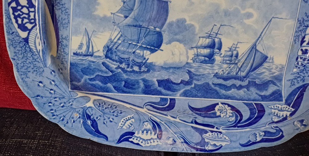 An oval blue and white earthenware transferware platter featuring a central naval sea battle scene surrounded by a botanical and seashell border. View X