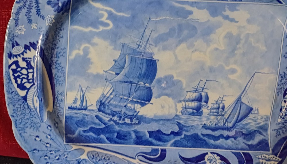 An oval blue and white earthenware transferware platter featuring a central naval sea battle scene surrounded by a botanical and seashell border. View XI