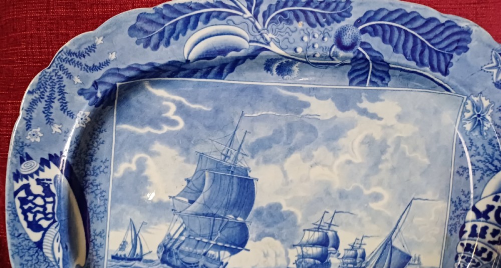 An oval blue and white earthenware transferware platter featuring a central naval sea battle scene surrounded by a botanical and seashell border. View XII