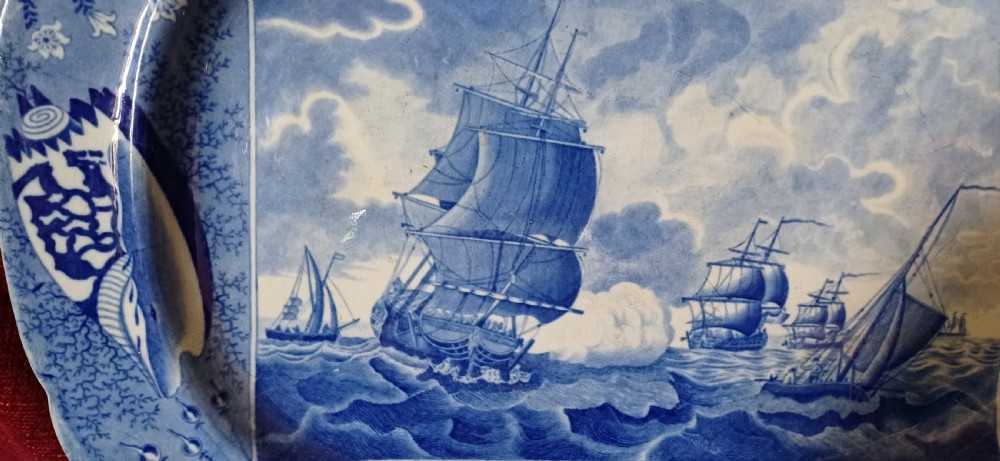 An oval blue and white earthenware transferware platter featuring a central naval sea battle scene surrounded by a botanical and seashell border. View XIII