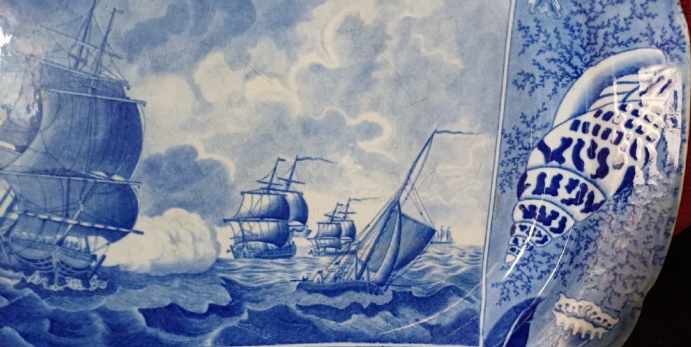 An oval blue and white earthenware transferware platter featuring a central naval sea battle scene surrounded by a botanical and seashell border. View XIV