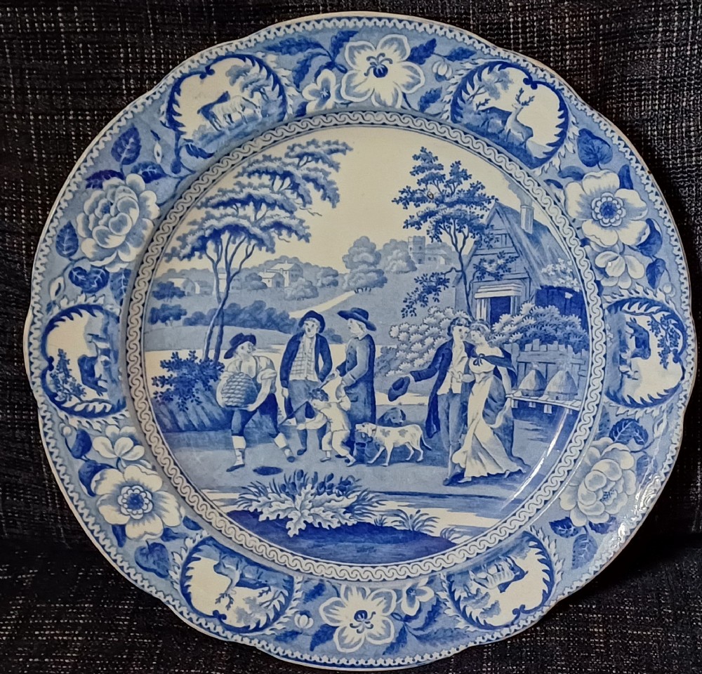 A circular blue and white transfer-printed earthenware plate featuring a central pastoral scene of figures near beehives surrounded by a floral and animal vignette border.