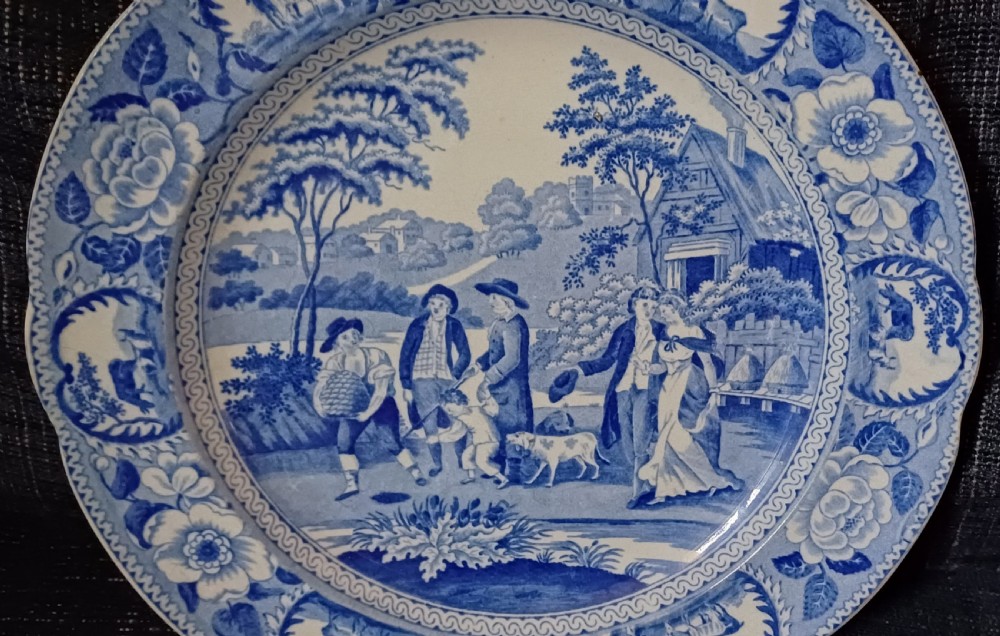 A circular blue and white transfer-printed earthenware plate featuring a central pastoral scene of figures near beehives surrounded by a floral and animal vignette border. View III