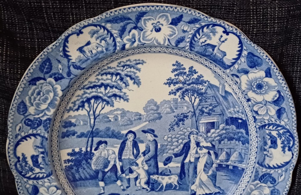 A circular blue and white transfer-printed earthenware plate featuring a central pastoral scene of figures near beehives surrounded by a floral and animal vignette border. View IV