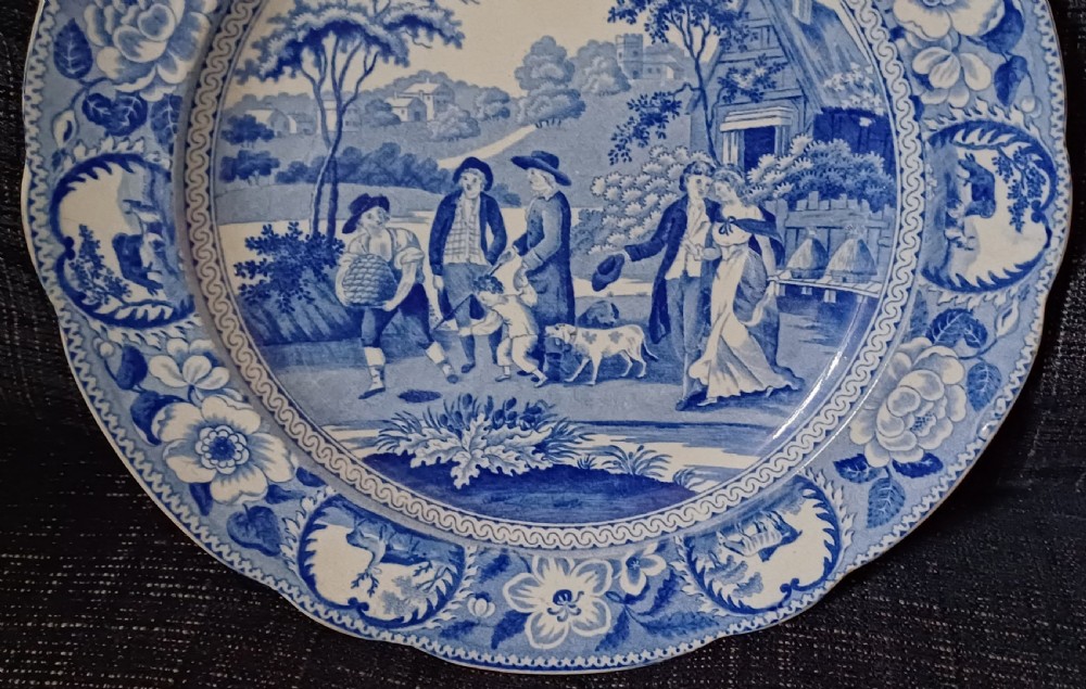 A circular blue and white transfer-printed earthenware plate featuring a central pastoral scene of figures near beehives surrounded by a floral and animal vignette border. View V