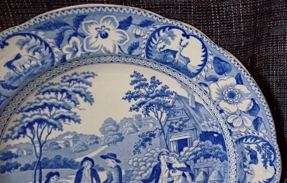 A circular blue and white transfer-printed earthenware plate featuring a central pastoral scene of figures near beehives surrounded by a floral and animal vignette border. View VI