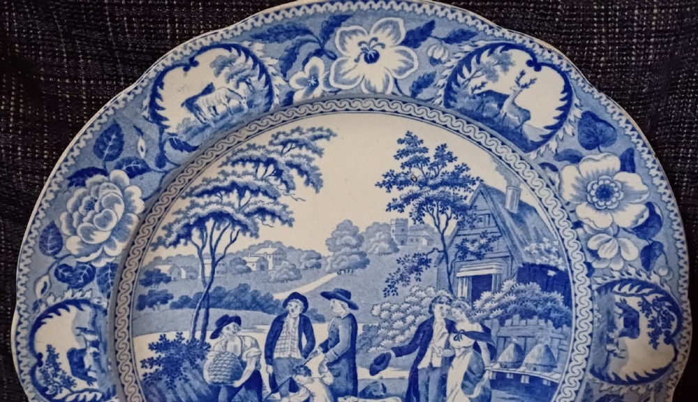 A circular blue and white transfer-printed earthenware plate featuring a central pastoral scene of figures near beehives surrounded by a floral and animal vignette border. View VII
