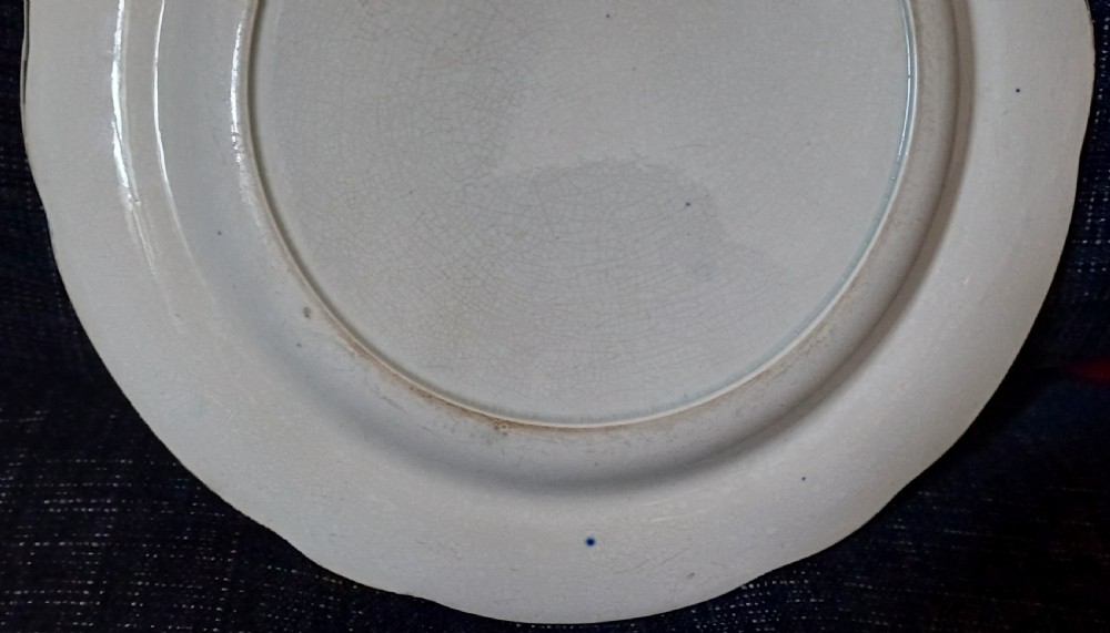 A circular blue and white transfer-printed earthenware plate featuring a central pastoral scene of figures near beehives surrounded by a floral and animal vignette border. View X