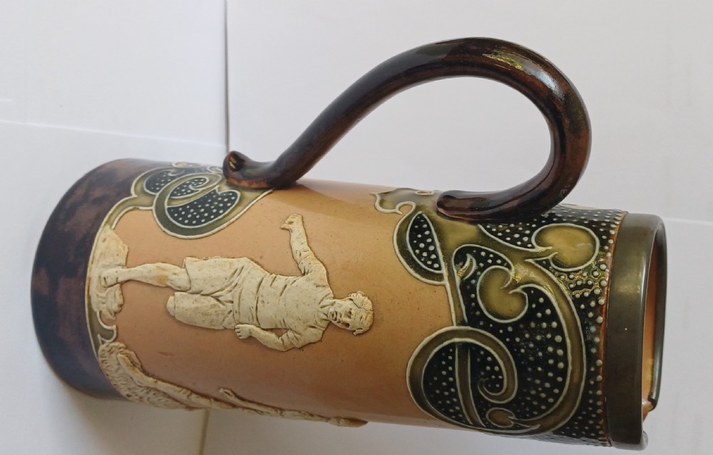 An Art Nouveau Doulton Lambeth glazed stoneware jug featuring relief figures of sports players alongside dark brown glazes and stylised swirl patterns. View XVII