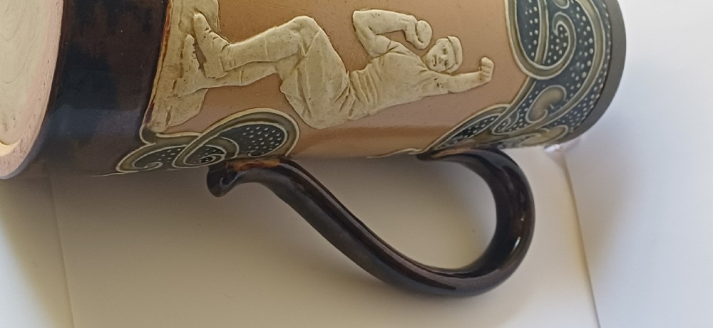 An Art Nouveau Doulton Lambeth glazed stoneware jug featuring relief figures of sports players alongside dark brown glazes and stylised swirl patterns. View XIX