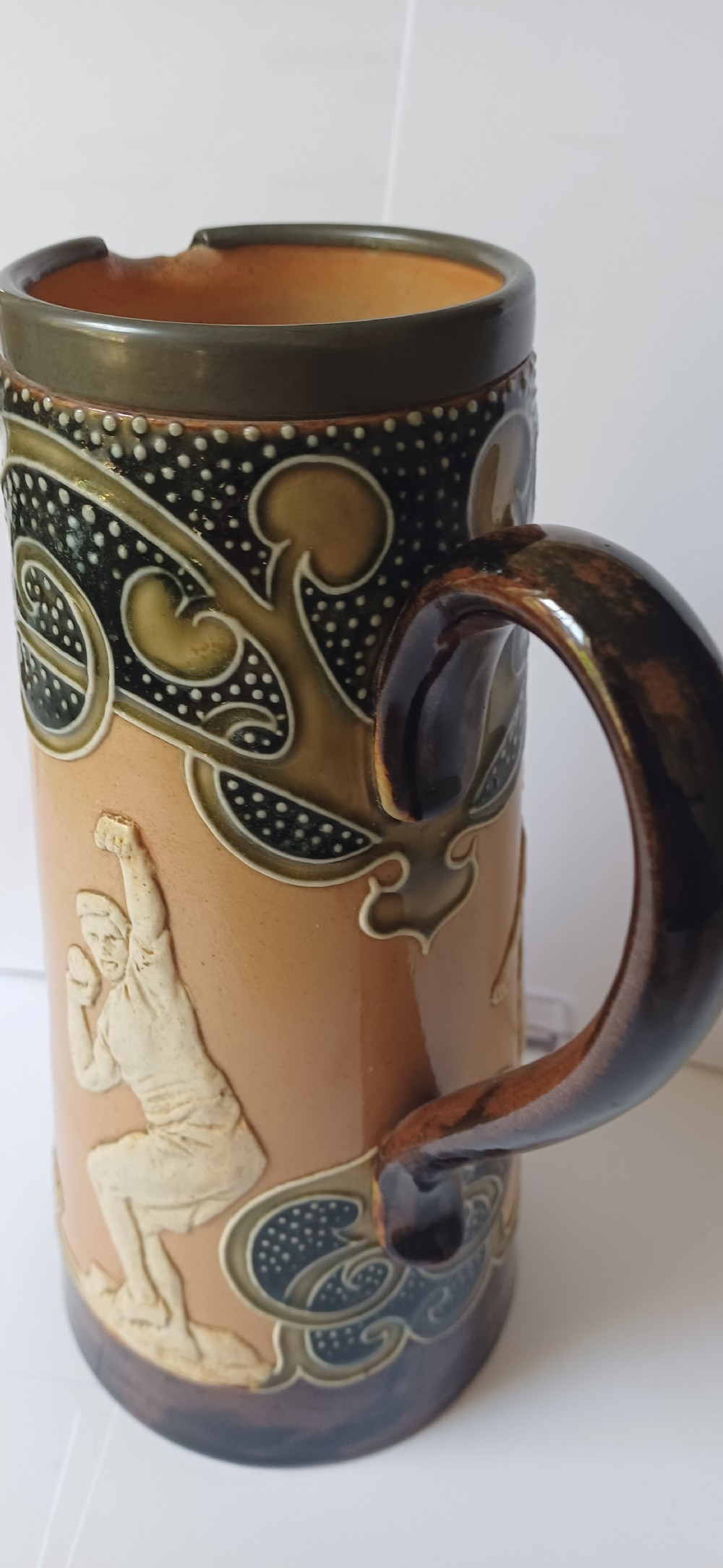 An Art Nouveau Doulton Lambeth glazed stoneware jug featuring relief figures of sports players alongside dark brown glazes and stylised swirl patterns. View XX