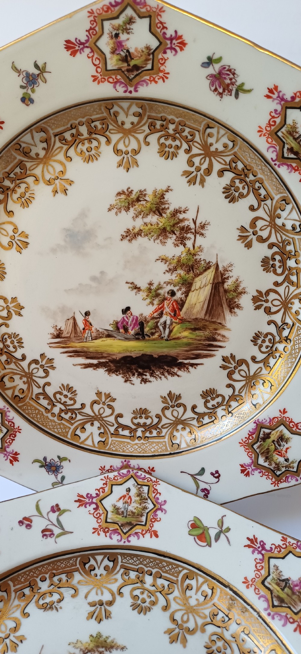 A pair of octagonal white porcelain cabinet plates featuring hand-painted romantic pastoral scenes in the centre, framed by elaborate gilt scrollwork, floral motifs, and decorative rim cartouches. View VIII