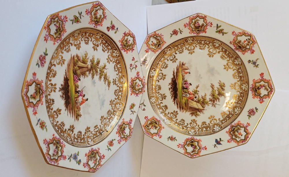 A pair of octagonal white porcelain cabinet plates featuring hand-painted romantic pastoral scenes in the centre, framed by elaborate gilt scrollwork, floral motifs, and decorative rim cartouches. View IX