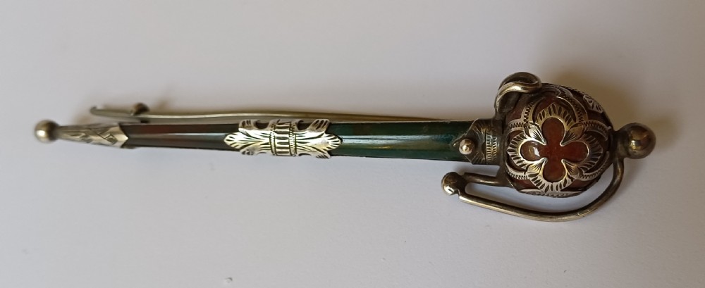 An antique silver kilt pin crafted in the shape of a Scottish basket-hilted broadsword inlaid with green and brown polished agate stones.