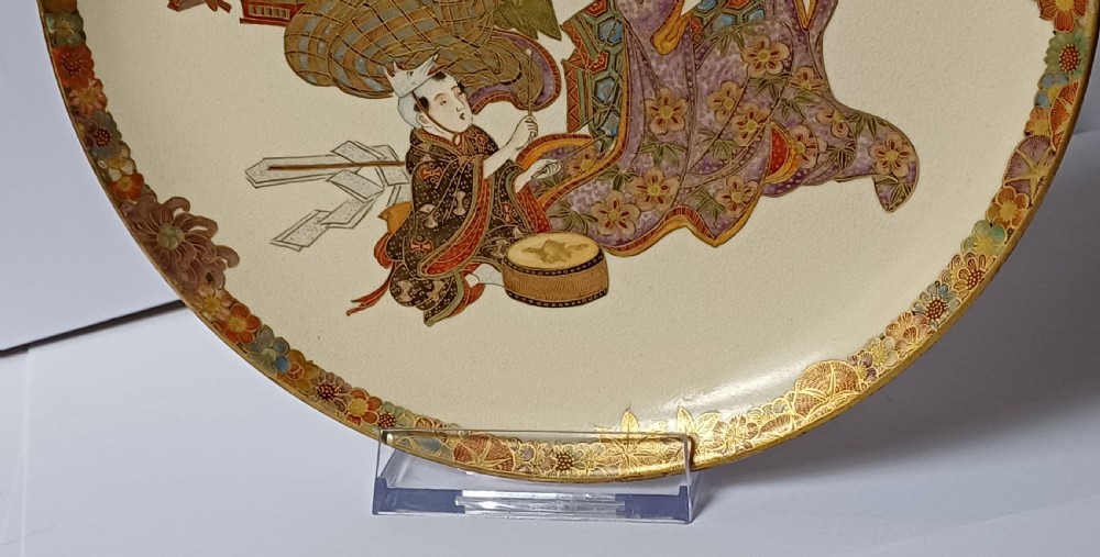 A circular shallow cream earthenware dish featuring hand-painted figures in traditional clothing framed by a gilded floral border. View XXII