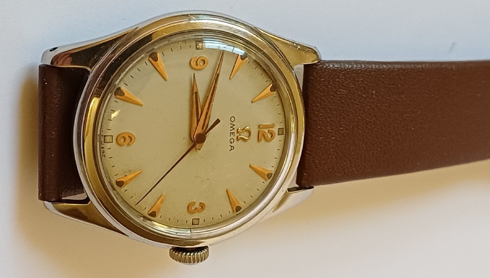 A vintage 1950s Omega wristwatch with a cream dial, gold hour markers, stainless steel case, and a smooth brown leather strap. View XII