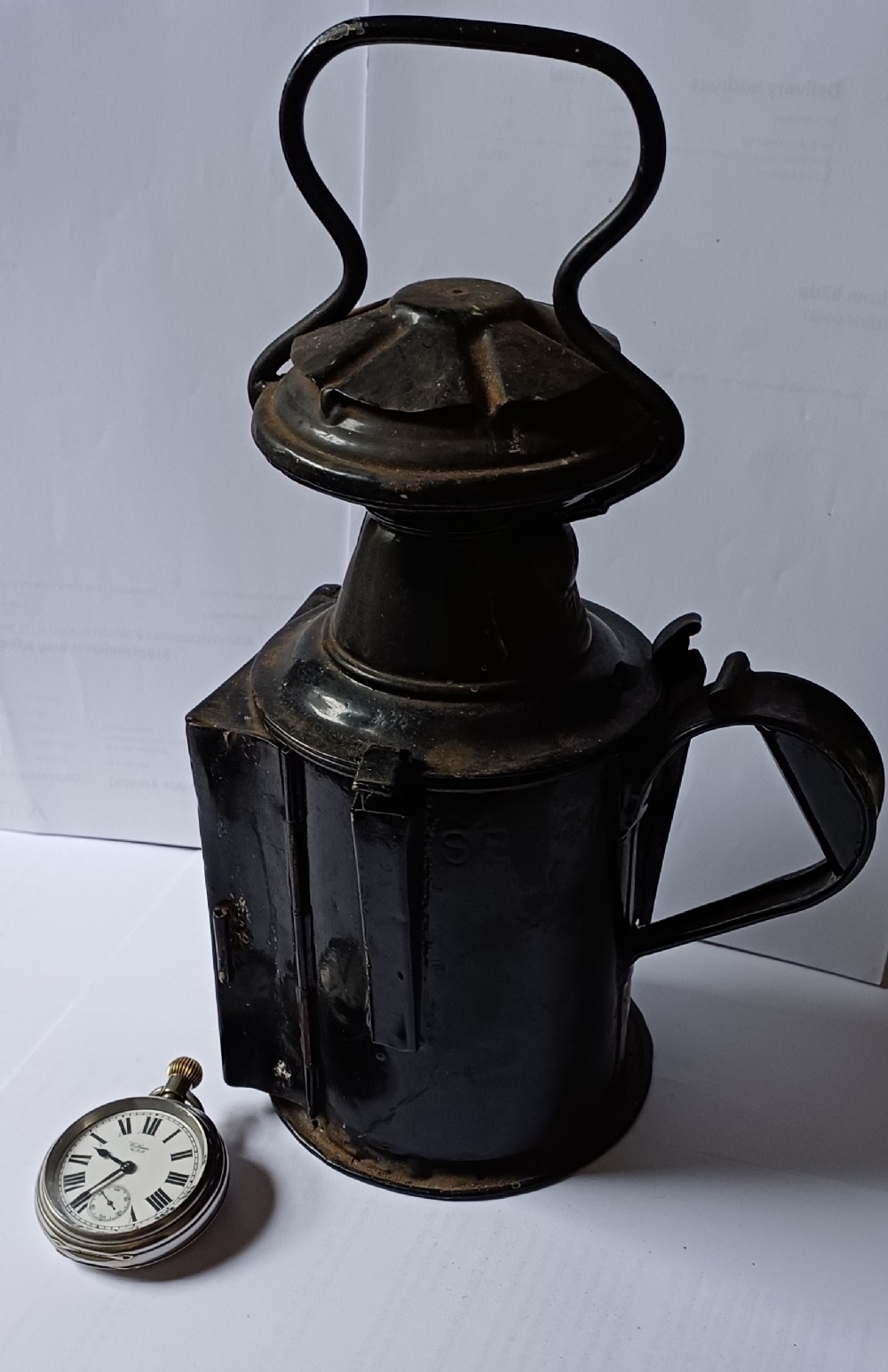 A black painted metal railway lantern with a top carrying handle alongside a silver-coloured pocket watch displaying Roman numerals.