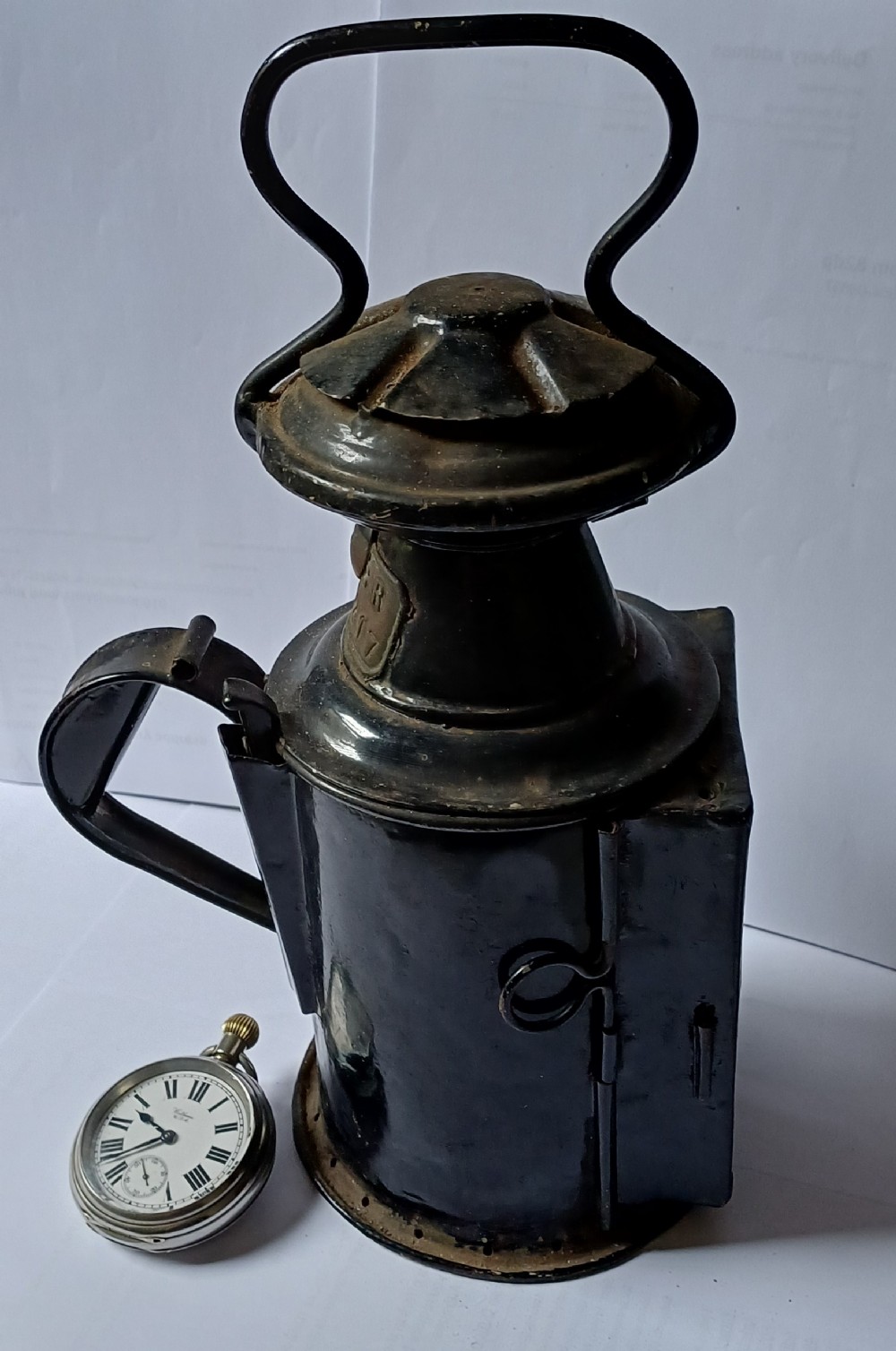 A black painted metal railway lantern with a top carrying handle alongside a silver-coloured pocket watch displaying Roman numerals. View II