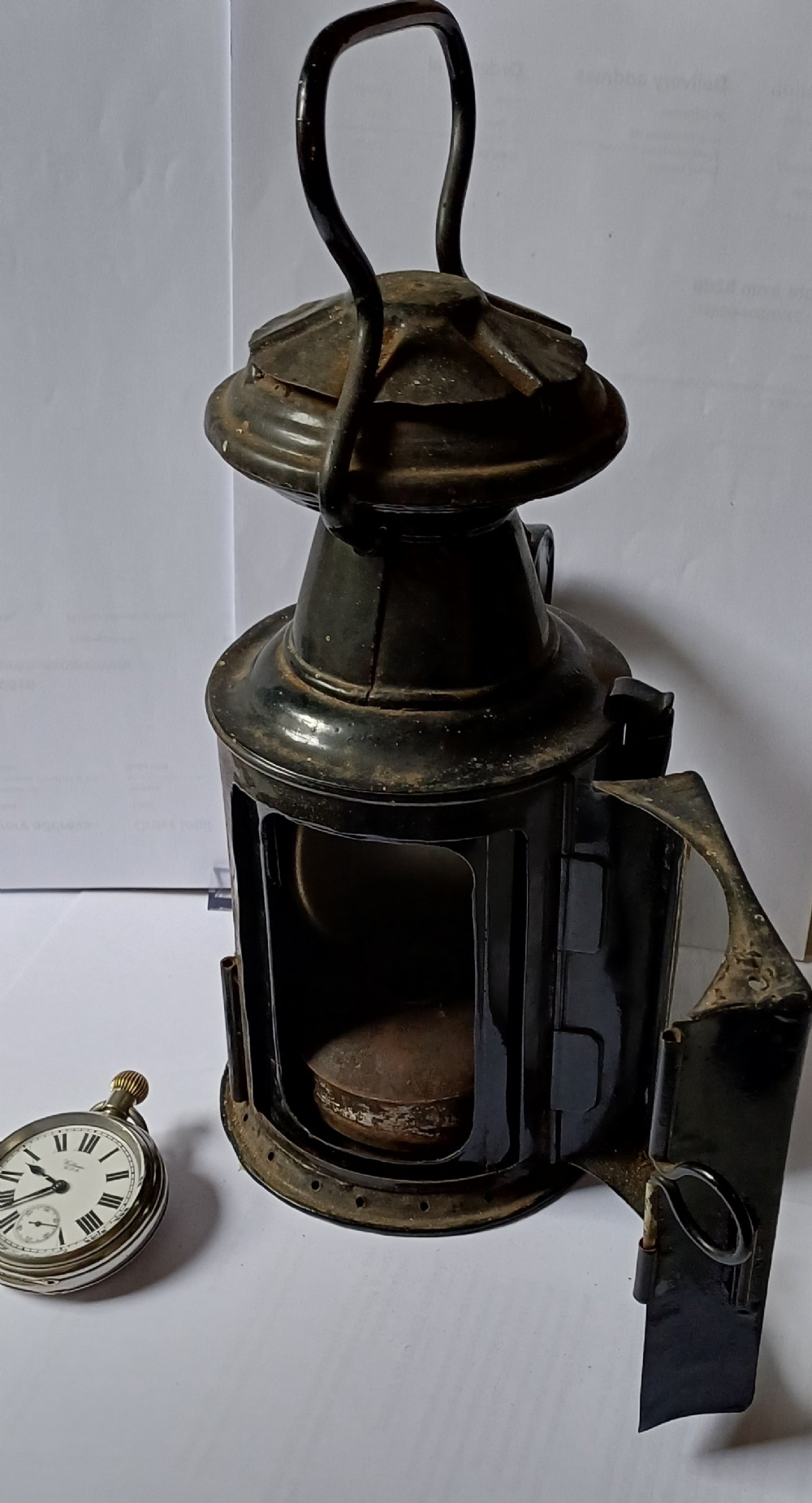 A black painted metal railway lantern with a top carrying handle alongside a silver-coloured pocket watch displaying Roman numerals. View III
