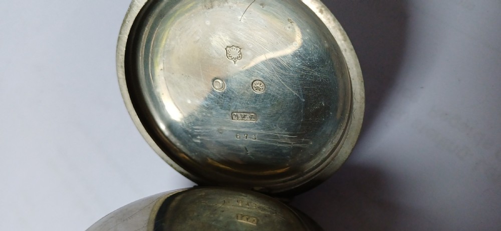 A black painted metal railway lantern with a top carrying handle alongside a silver-coloured pocket watch displaying Roman numerals. View XIV