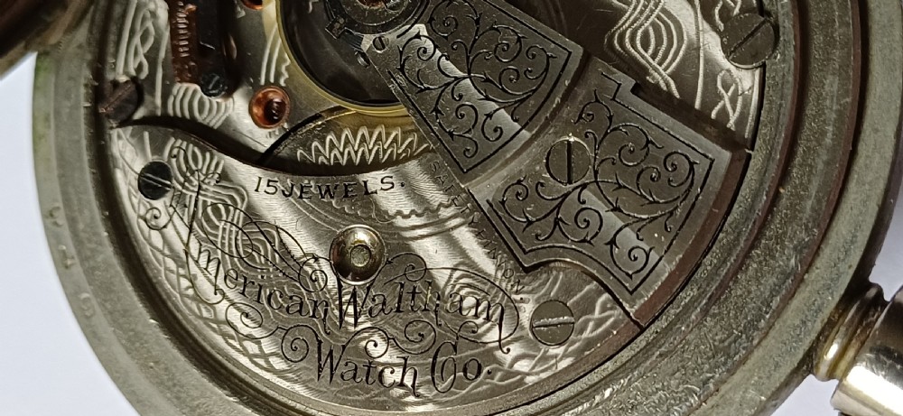 A black painted metal railway lantern with a top carrying handle alongside a silver-coloured pocket watch displaying Roman numerals. View XX