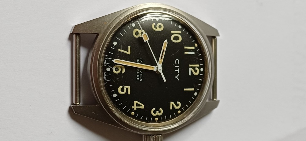 A vintage mid-century military wristwatch featuring a round steel case, a black dial with luminous Arabic numerals, outer minute markings, and luminescent hands. View II
