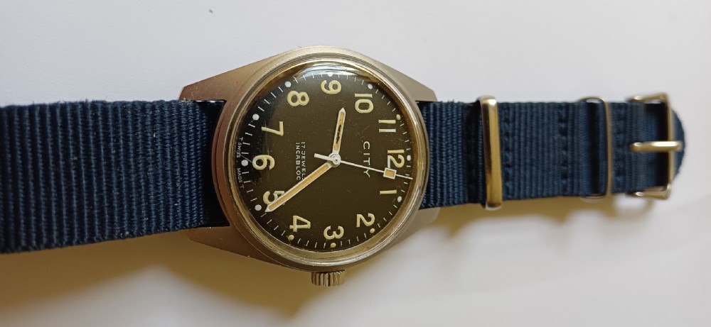 A vintage mid-century military wristwatch featuring a round steel case, a black dial with luminous Arabic numerals, outer minute markings, and luminescent hands. View III
