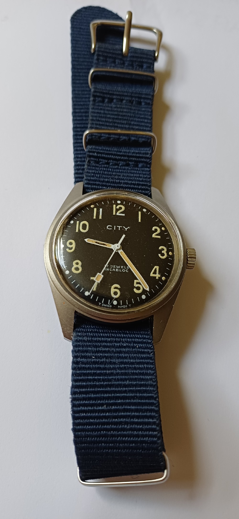 A vintage mid-century military wristwatch featuring a round steel case, a black dial with luminous Arabic numerals, outer minute markings, and luminescent hands. View IV