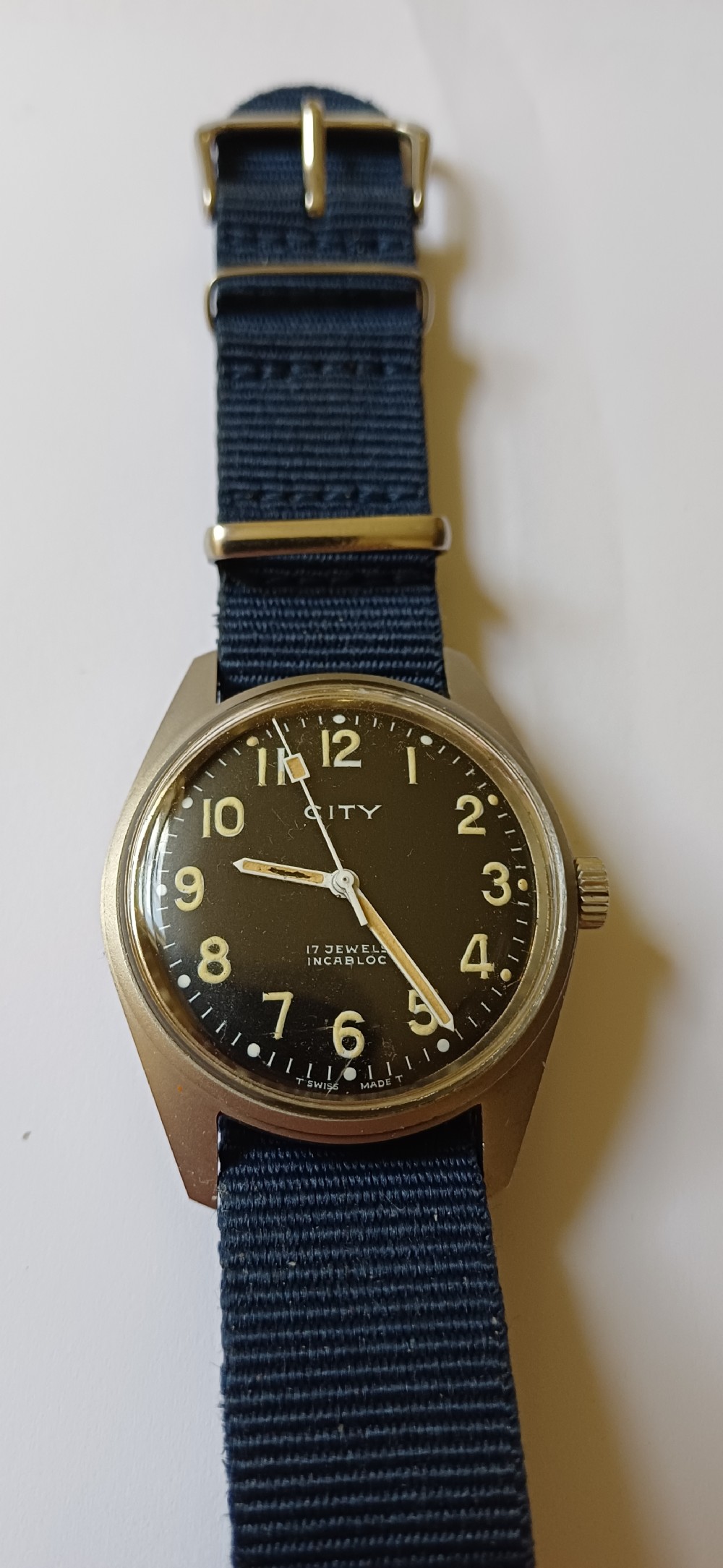 A vintage mid-century military wristwatch featuring a round steel case, a black dial with luminous Arabic numerals, outer minute markings, and luminescent hands. View V