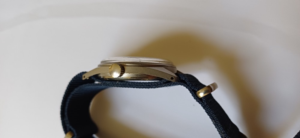 A vintage mid-century military wristwatch featuring a round steel case, a black dial with luminous Arabic numerals, outer minute markings, and luminescent hands. View VI