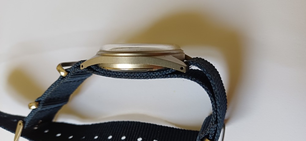 A vintage mid-century military wristwatch featuring a round steel case, a black dial with luminous Arabic numerals, outer minute markings, and luminescent hands. View VII