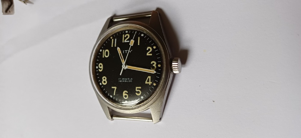 A vintage mid-century military wristwatch featuring a round steel case, a black dial with luminous Arabic numerals, outer minute markings, and luminescent hands. View XVI