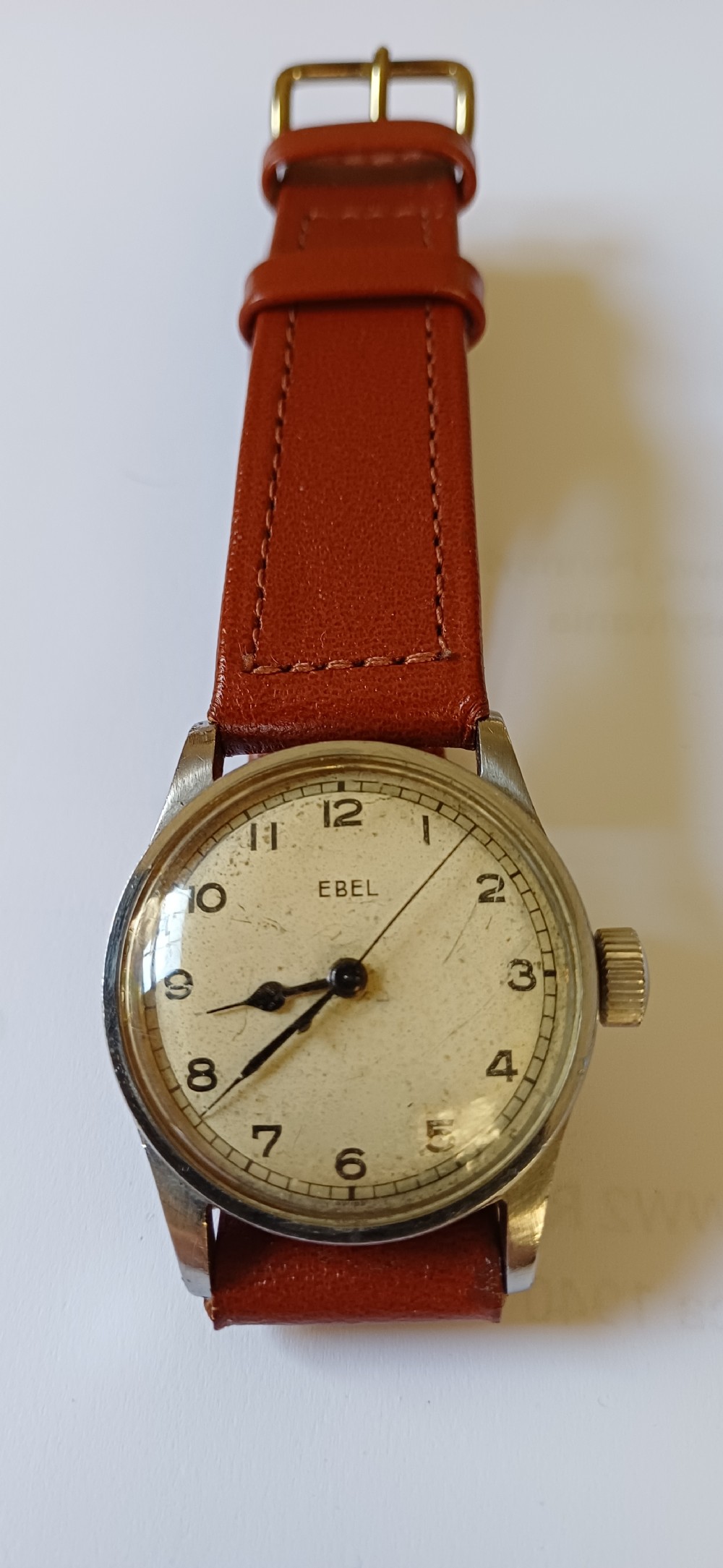 A vintage circular wristwatch featuring a cream dial with black Arabic numerals, blued steel hands, a metal case, and a brown leather strap.