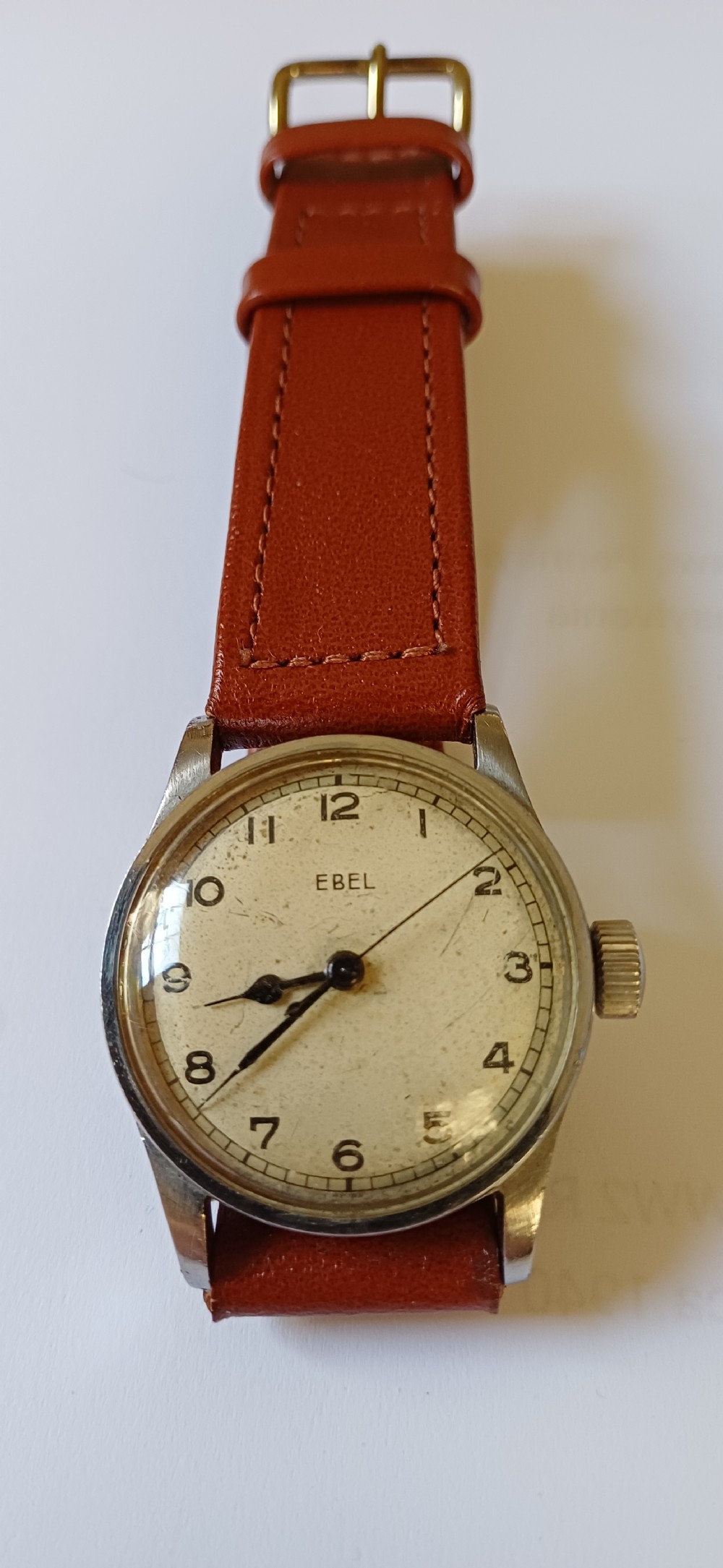 A vintage circular wristwatch featuring a cream dial with black Arabic numerals, blued steel hands, a metal case, and a brown leather strap. View II