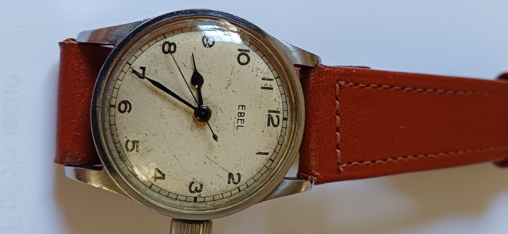 A vintage circular wristwatch featuring a cream dial with black Arabic numerals, blued steel hands, a metal case, and a brown leather strap. View III