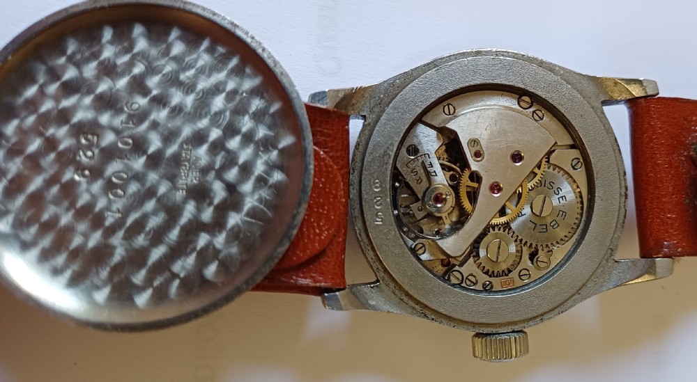 A vintage circular wristwatch featuring a cream dial with black Arabic numerals, blued steel hands, a metal case, and a brown leather strap. View XI