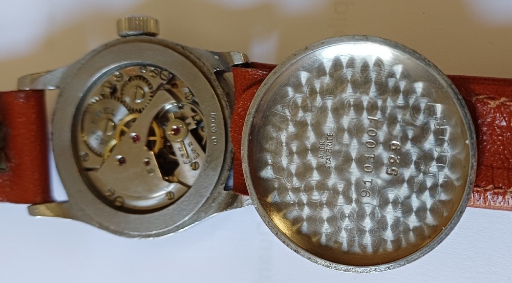 A vintage circular wristwatch featuring a cream dial with black Arabic numerals, blued steel hands, a metal case, and a brown leather strap. View XII