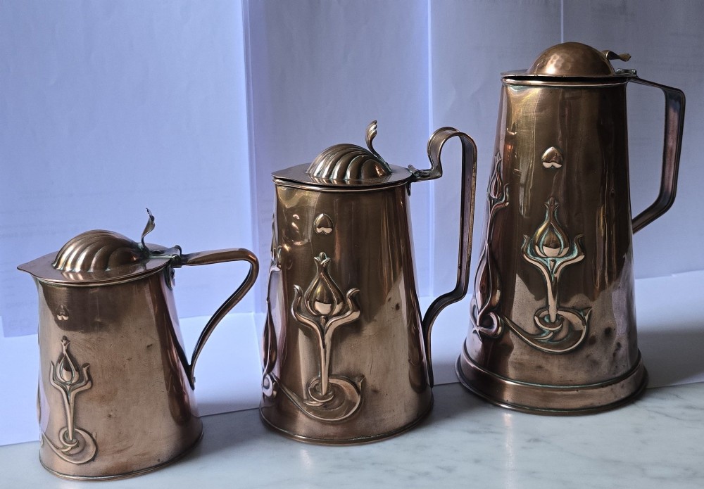 A set of three graduated copper jugs featuring embossed Art Nouveau floral motifs and shell-shaped hinged lids.