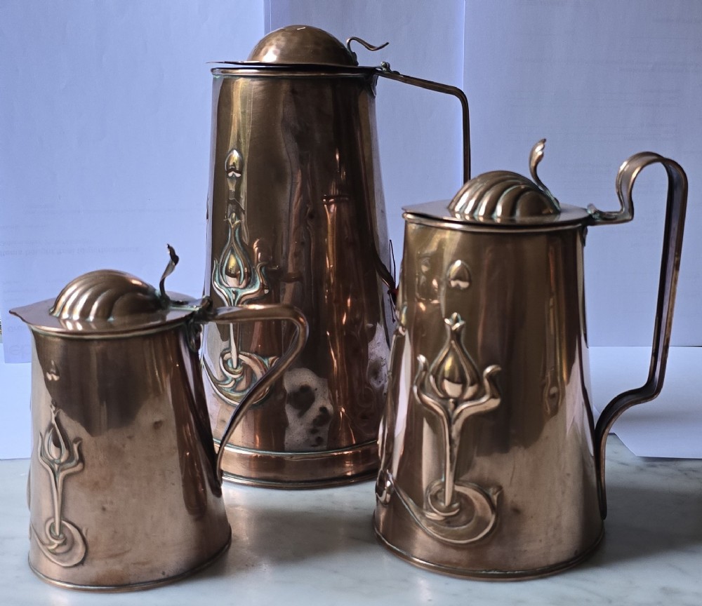 A set of three graduated copper jugs featuring embossed Art Nouveau floral motifs and shell-shaped hinged lids. View II