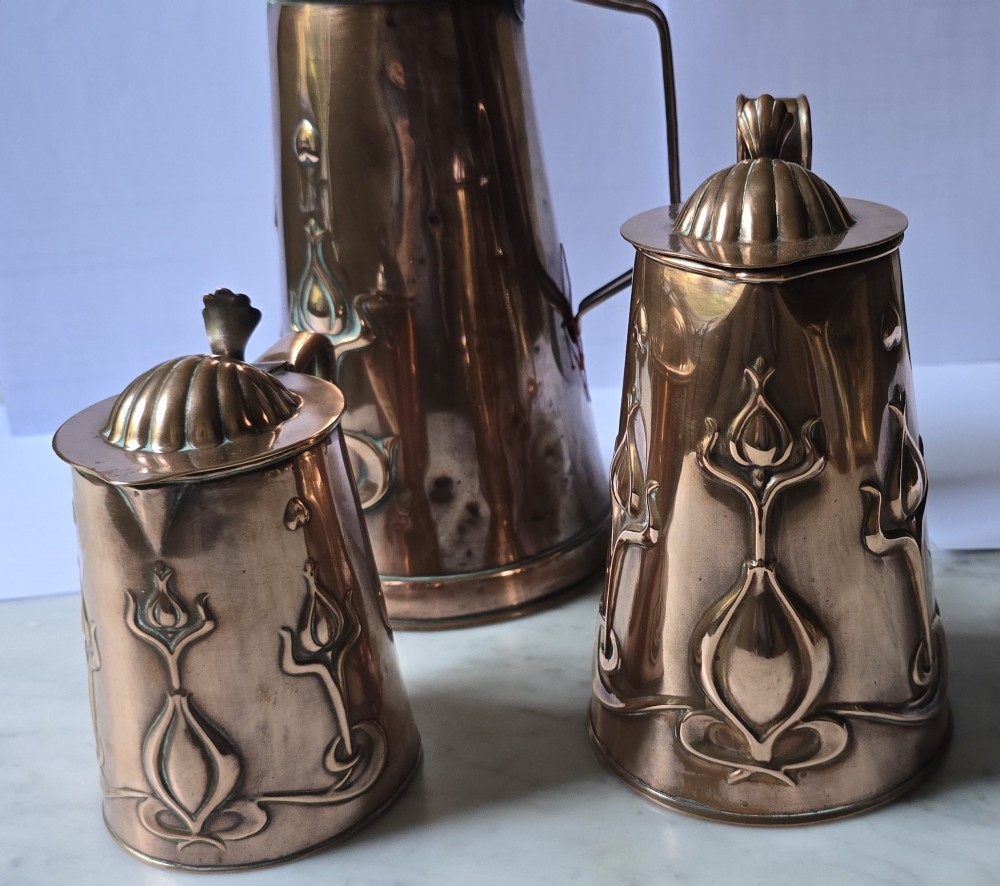 A set of three graduated copper jugs featuring embossed Art Nouveau floral motifs and shell-shaped hinged lids. View V