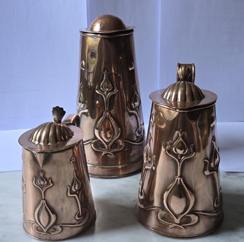 A set of three graduated copper jugs featuring embossed Art Nouveau floral motifs and shell-shaped hinged lids. View VI